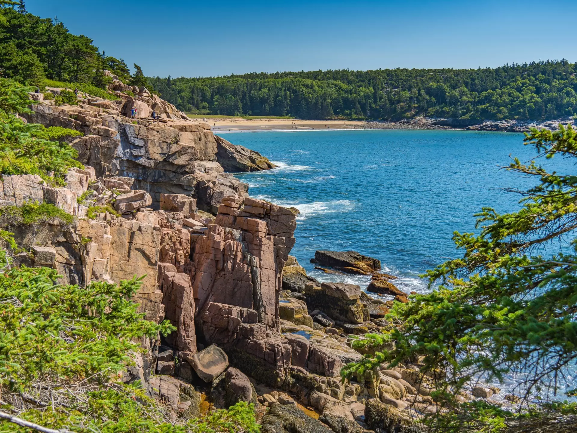 A rocky coastline covered in dense forest with an arc of golden sand on a distant beach.
