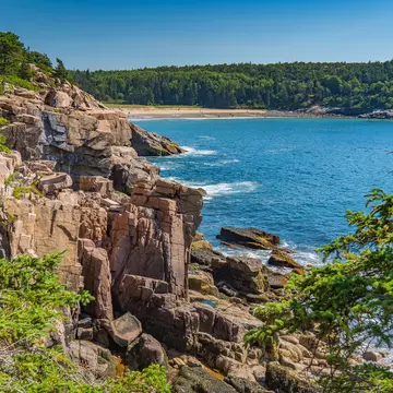 A rocky coastline covered in dense forest with an arc of golden sand on a distant beach.