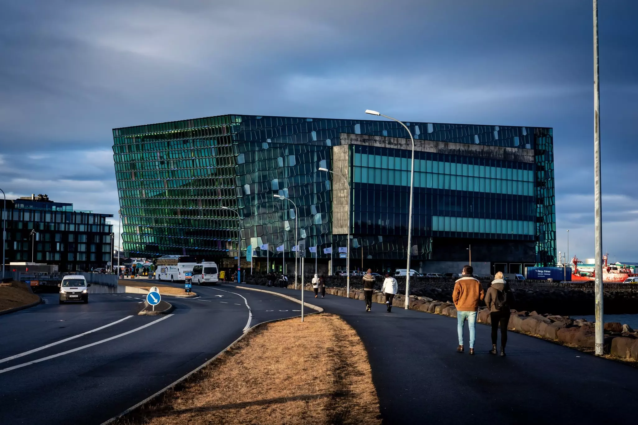People walk alongside cars on a road leading to a modern glass building in a city.