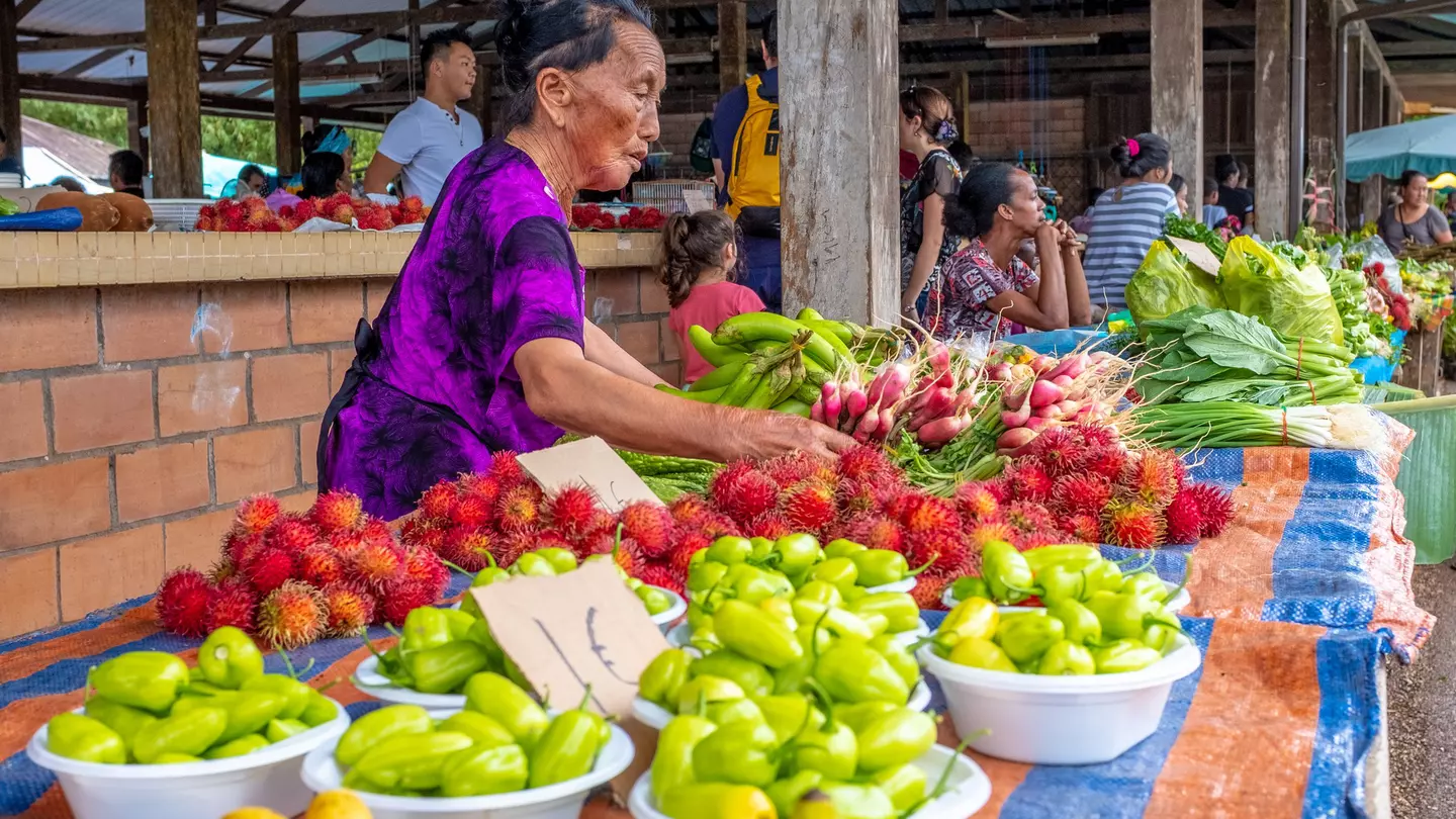 A few local people sit beneath the white stone pillars of Taukkyan War Cemetery's central monument. In the foreground numerous graves, with small headstones, are visible surrounded by manicured lawns.