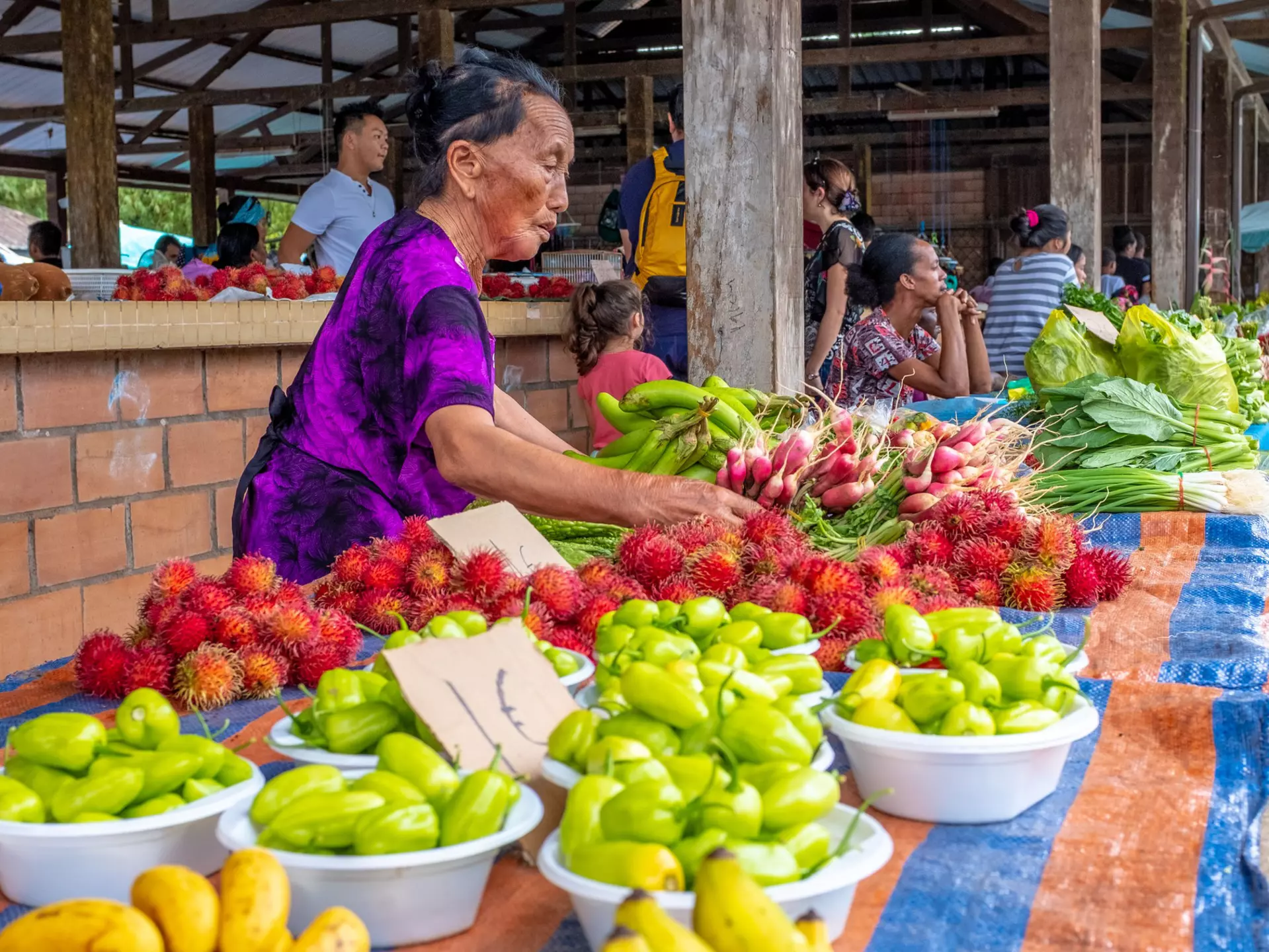 A few local people sit beneath the white stone pillars of Taukkyan War Cemetery's central monument. In the foreground numerous graves, with small headstones, are visible surrounded by manicured lawns.