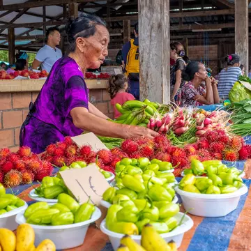A few local people sit beneath the white stone pillars of Taukkyan War Cemetery's central monument. In the foreground numerous graves, with small headstones, are visible surrounded by manicured lawns.