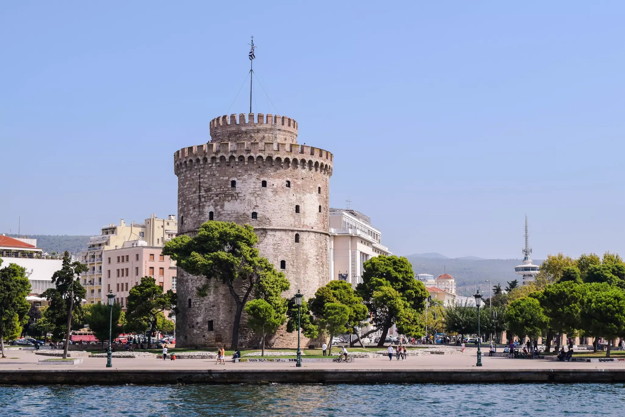 A large round fortified structure surrounded by trees is seen along the waterfront of a city.
