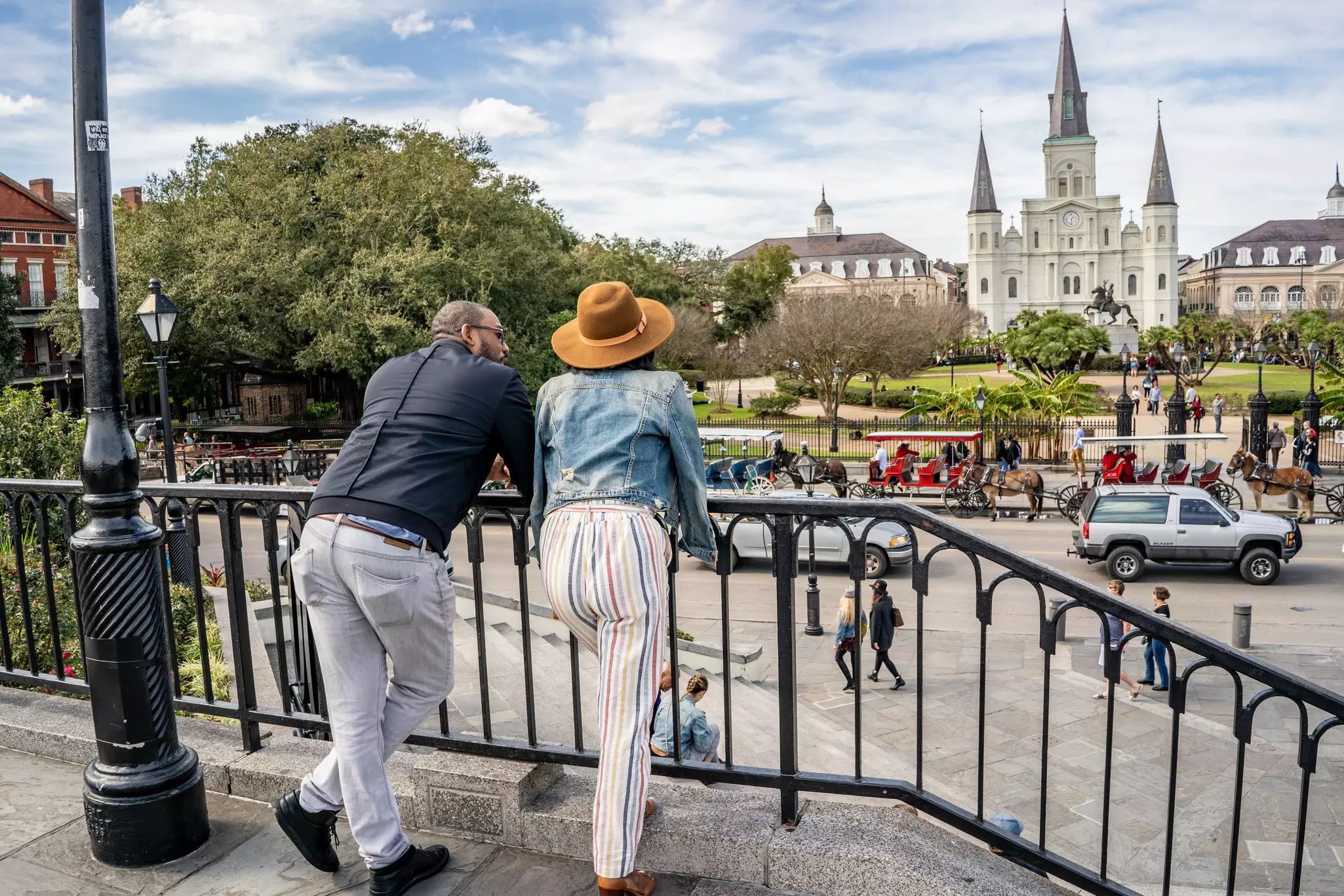 Two people are seen from behind leaning on a wrought-iron fence and looking out a city square with people and vehicles. A church is visible at the far end of the square.