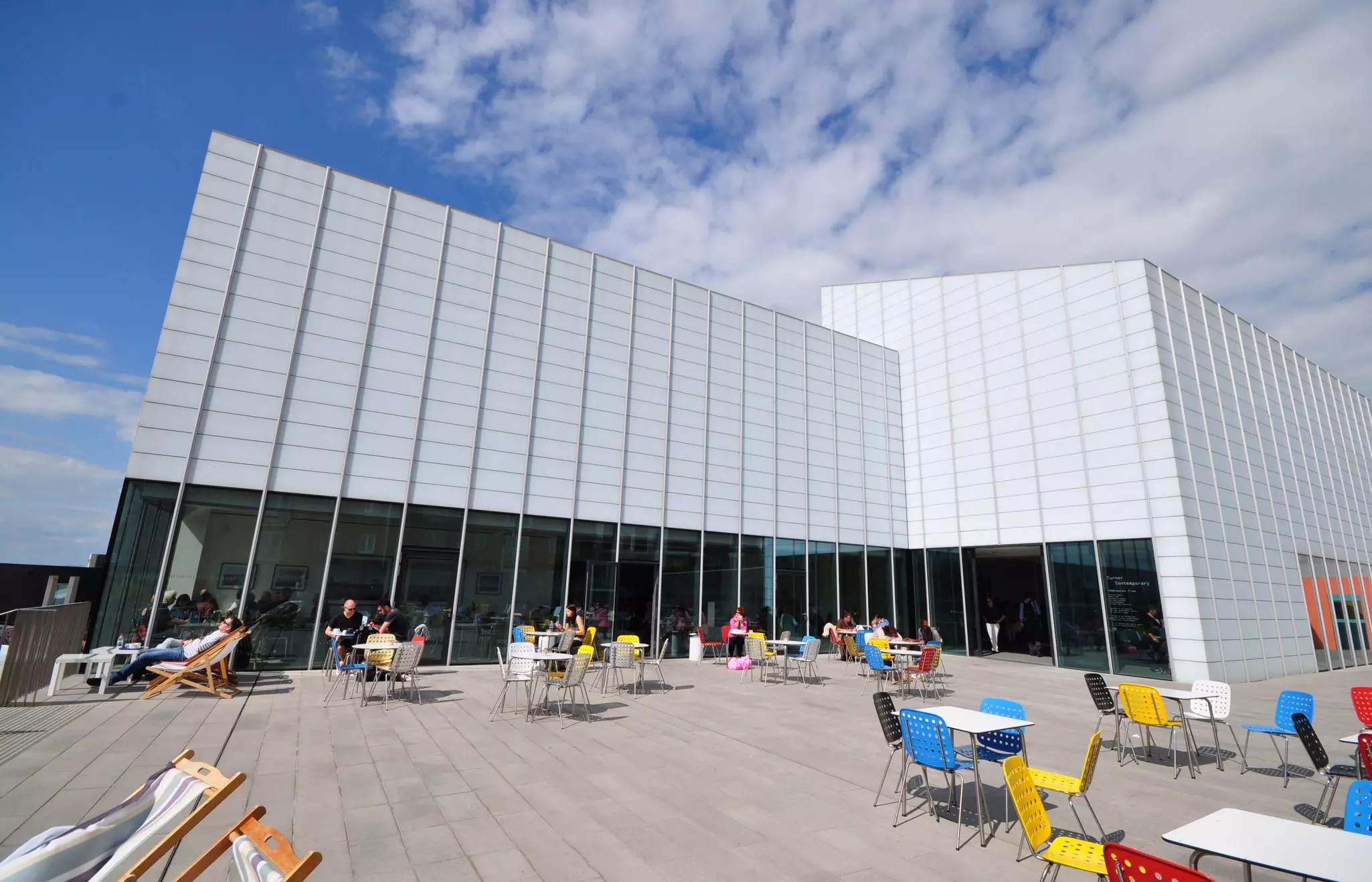 Exterior of modern art museum with people sitting at outdoor patio tables with colorful chairs on a sunny day.