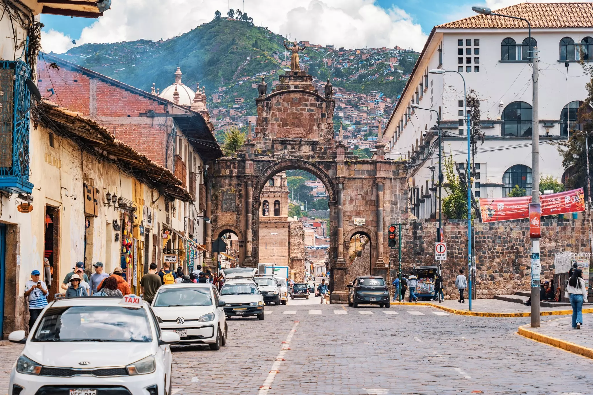 Cars drive through a stone archway in a historic city by a hillside.