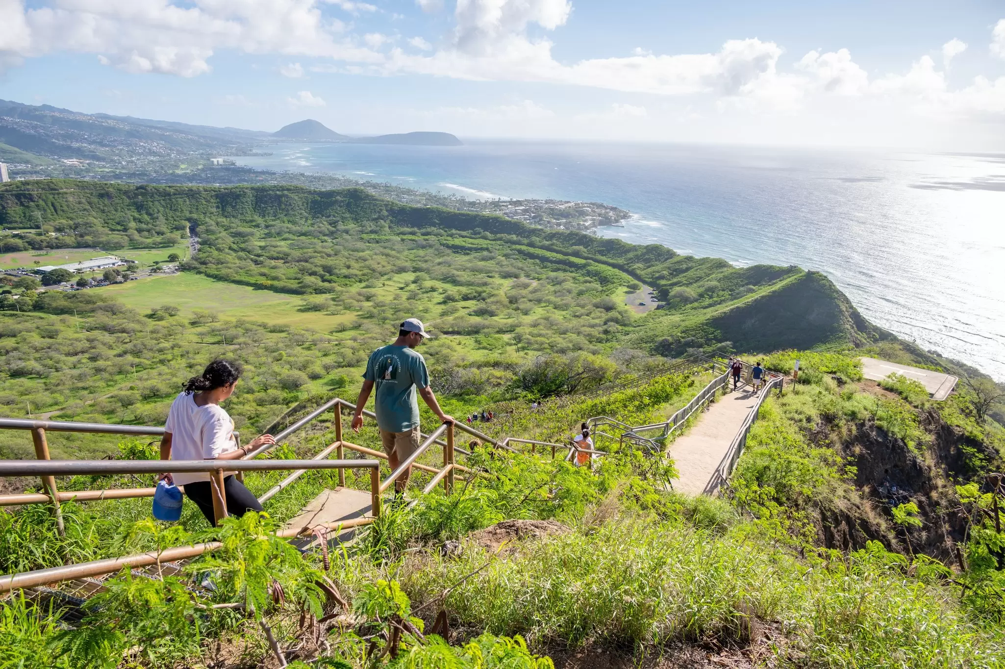 Hikers follow a cliff-top trail through folling green fields down towards a headland; Diamond Head lookout trail, Hawaii.