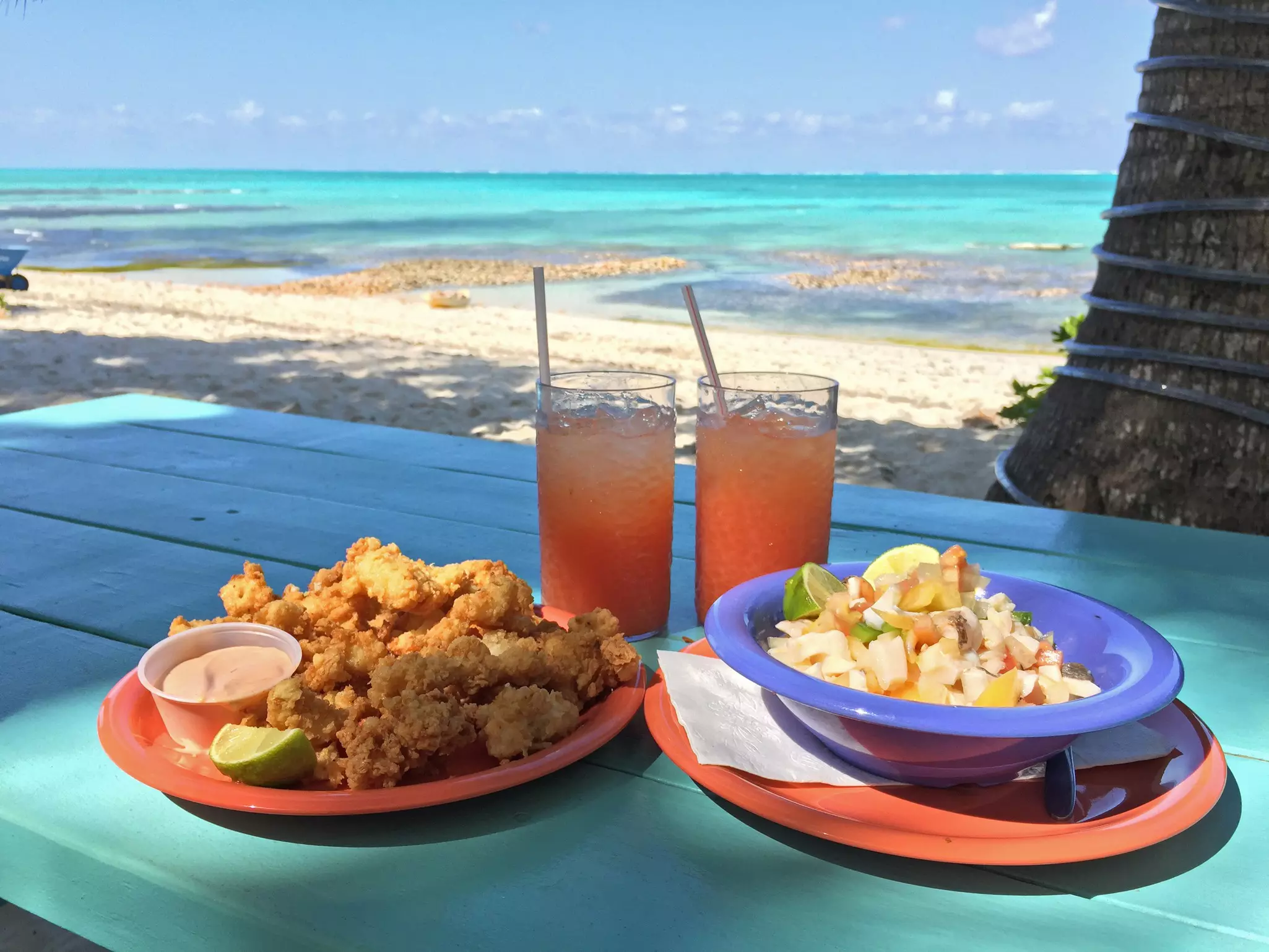 Two plates and two drinks on a turquoise table near the beach. One plate has cracked conch and the other has conch salad. Both drinks are bright coral color and have ice and straws in them.