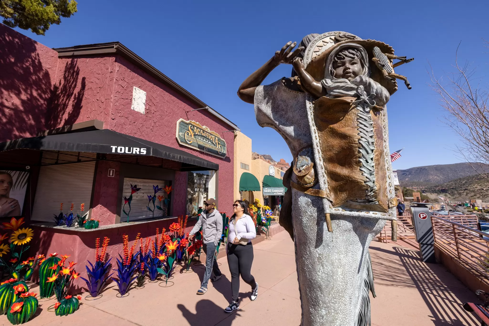An outdoor sculpture of a child on an adult's back is installed on a sidewalk in front of stores.