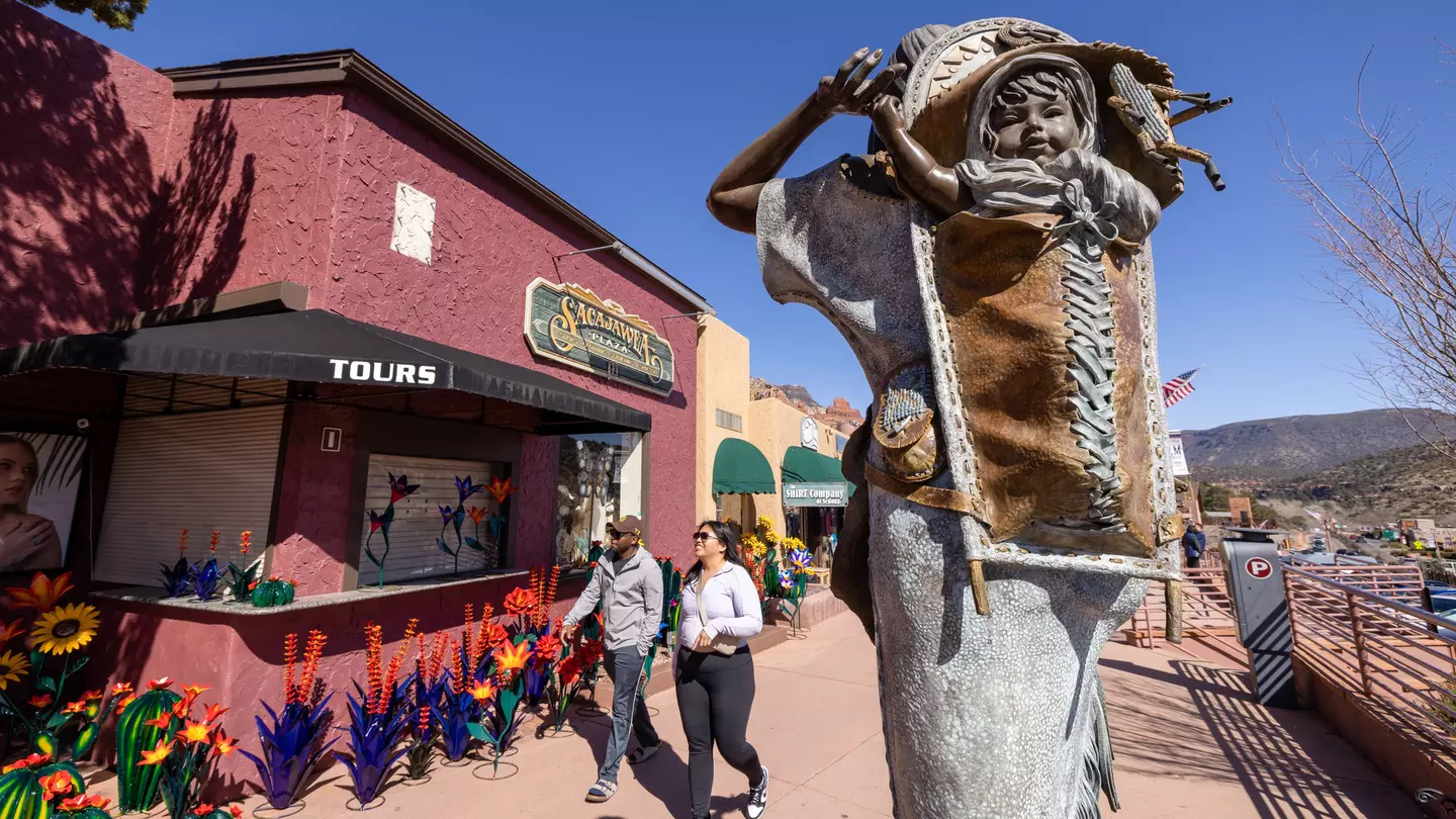 An outdoor sculpture of a child on an adult's back is installed on a sidewalk in front of stores.