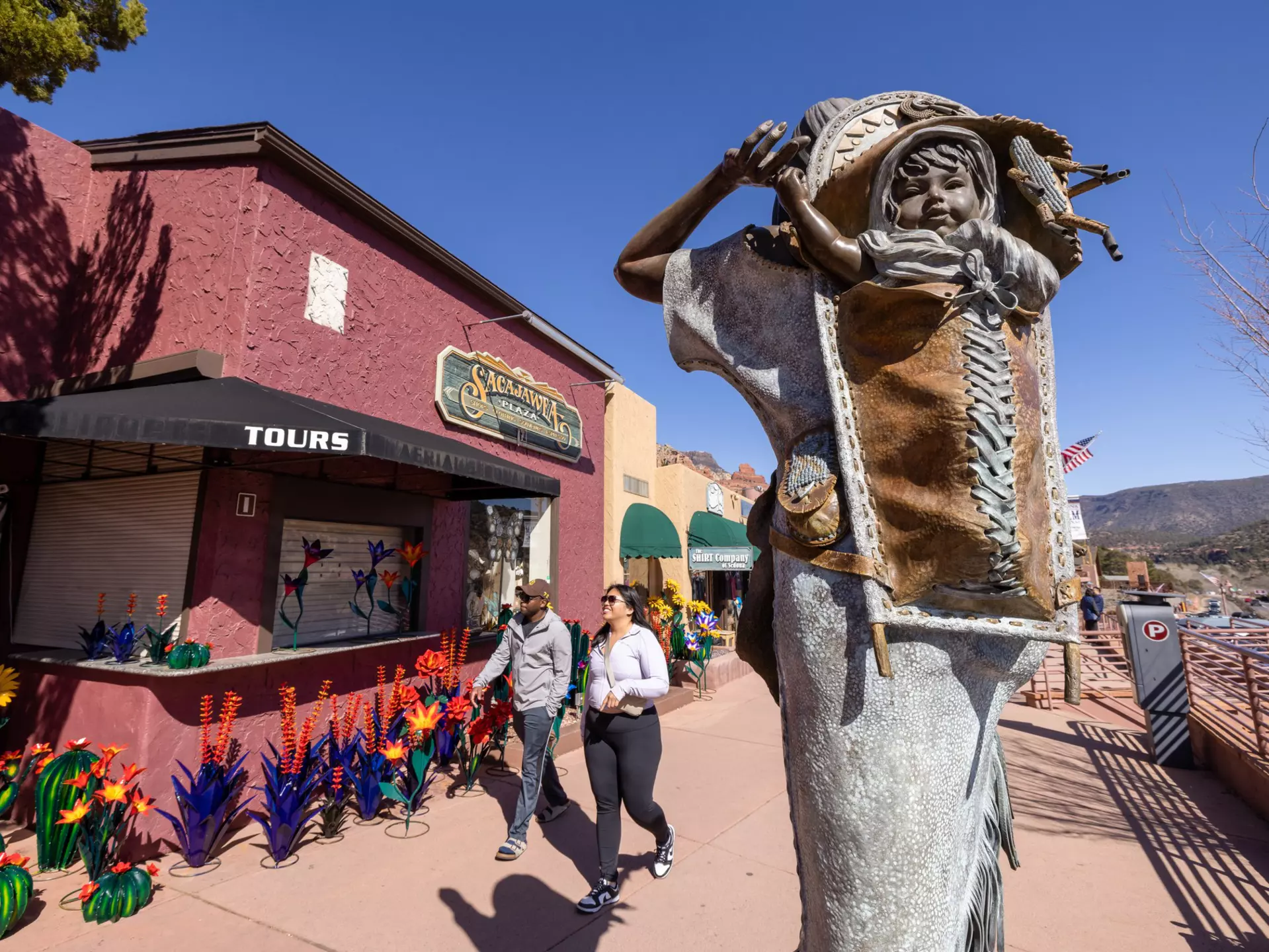 An outdoor sculpture of a child on an adult's back is installed on a sidewalk in front of stores.