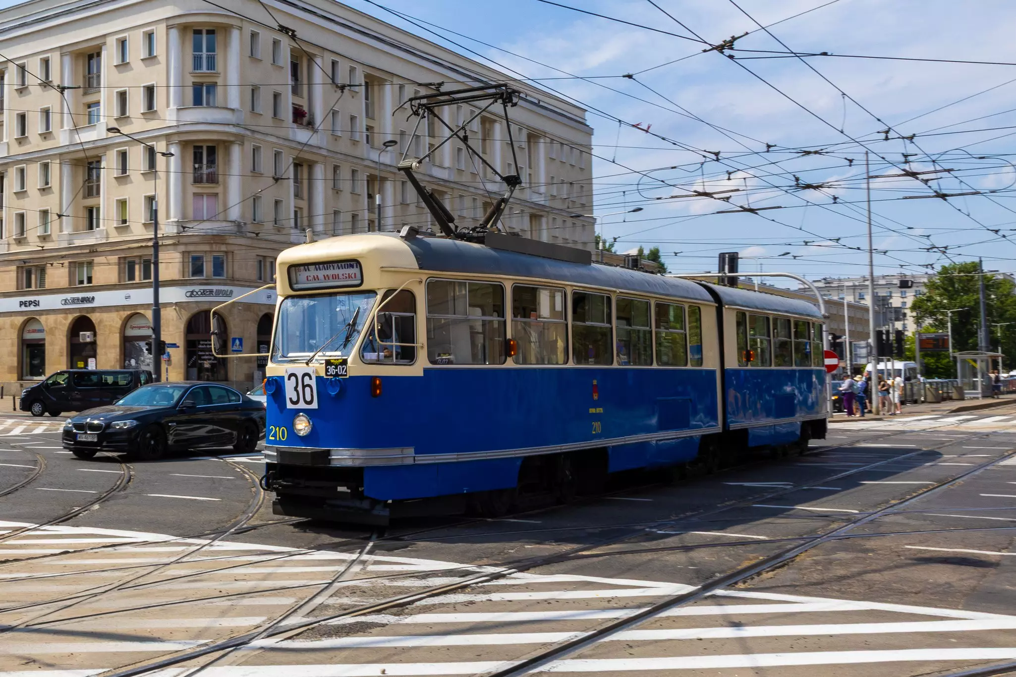 A tram with a blue body and a cream-colored roof passes through an intersection crisscrossed with tram wires in Warsaw, Poland.