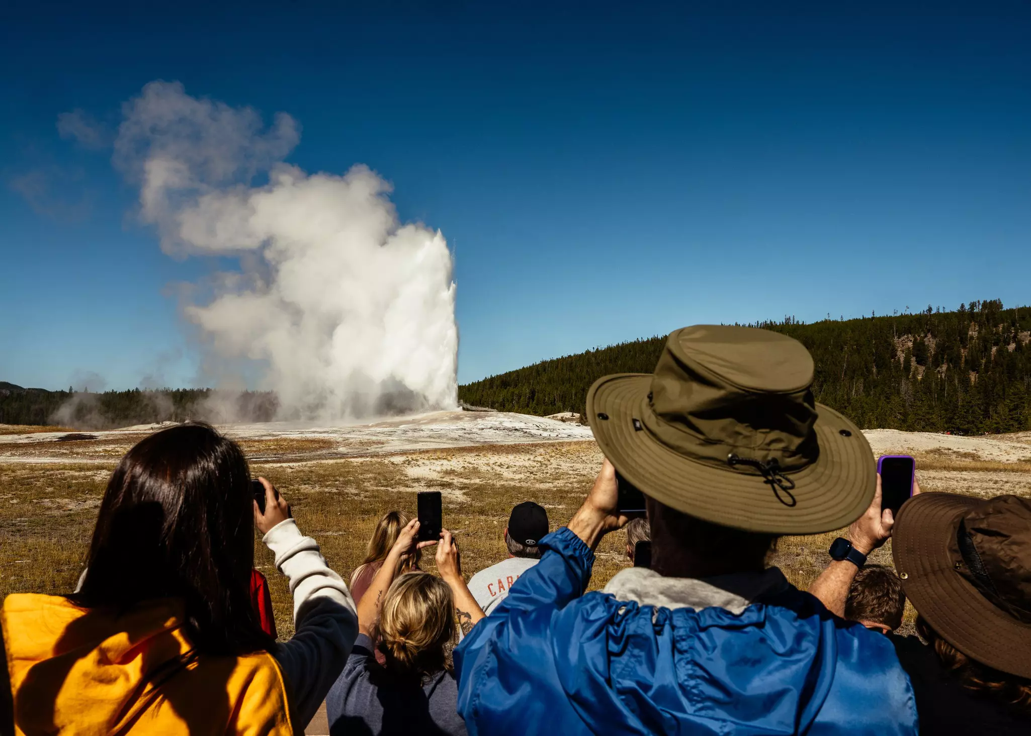 Tourists all stand facing a geyser as it shoots into the air. All of them hold their phones up to take a photo of the natural wonder.