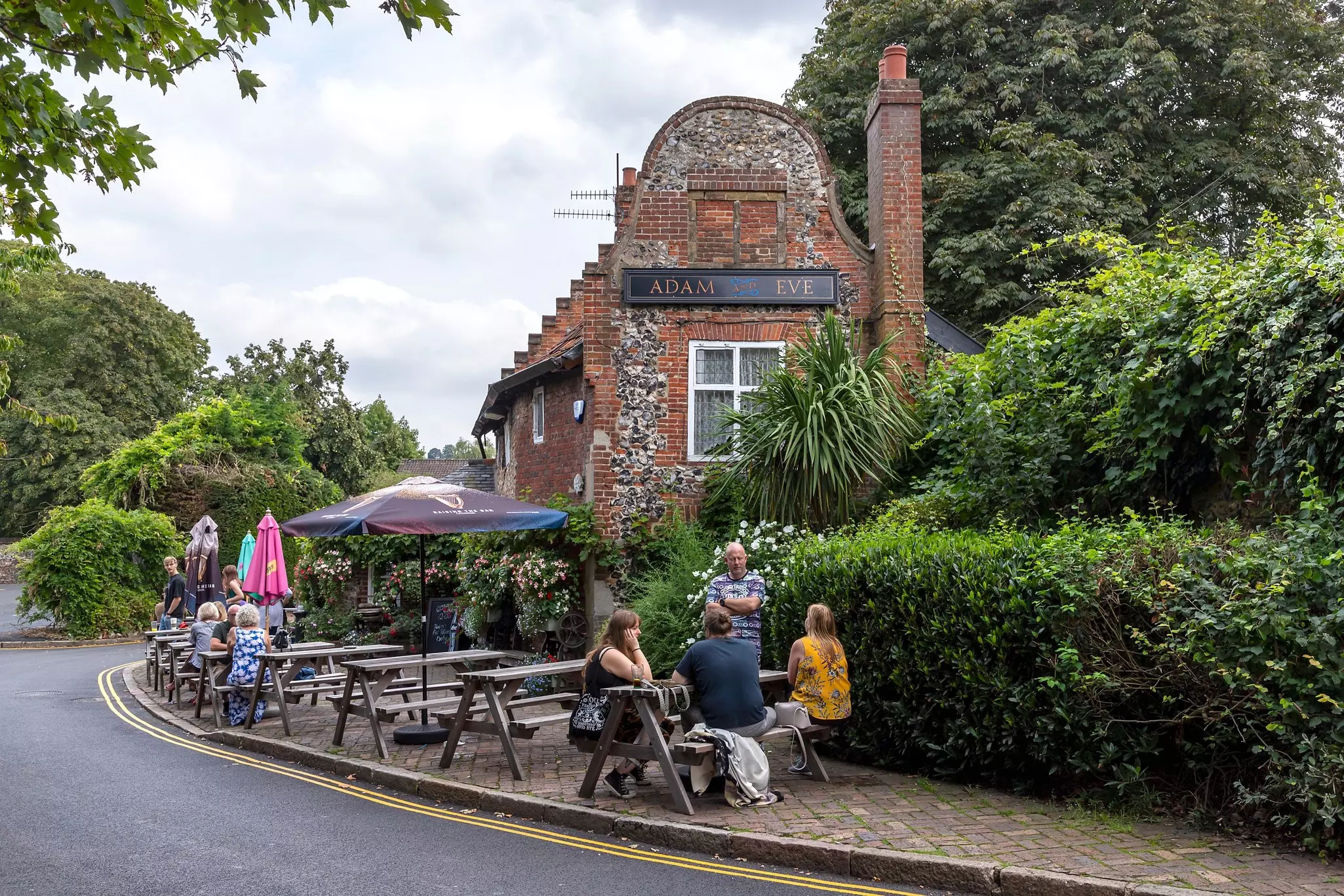 Patrons sit at picnic tables on a sidewalk outside a pub on a leafy town street.
