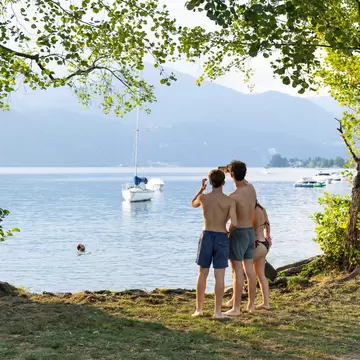 Three people in swimwear under leafy trees at a grassy lakeshore, looking out at a moored sailboat and hazy hills