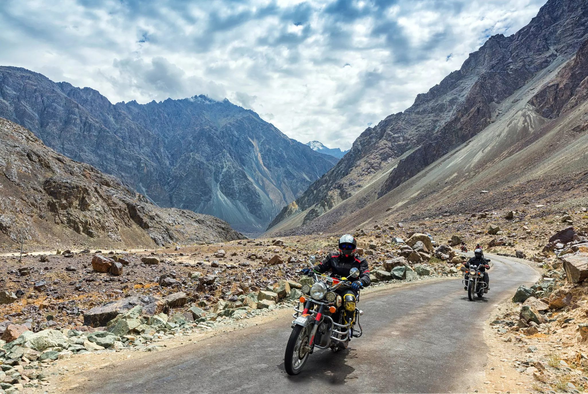 Motorcyclists drive along a narrow paved roads in a rocky mountainous region
