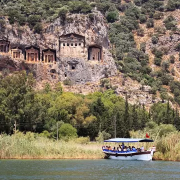 Temple-fronted tombs carved into a rocky cliff above green trees and reeds, with a canopied tour boat on the river below.