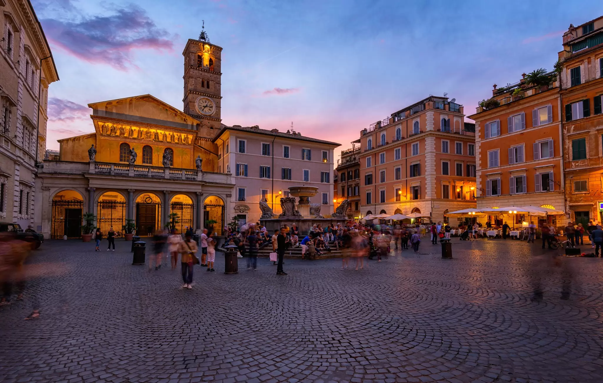 A church on a plaza with a fountain at sunset; people are seated around the fountain and at cafes.