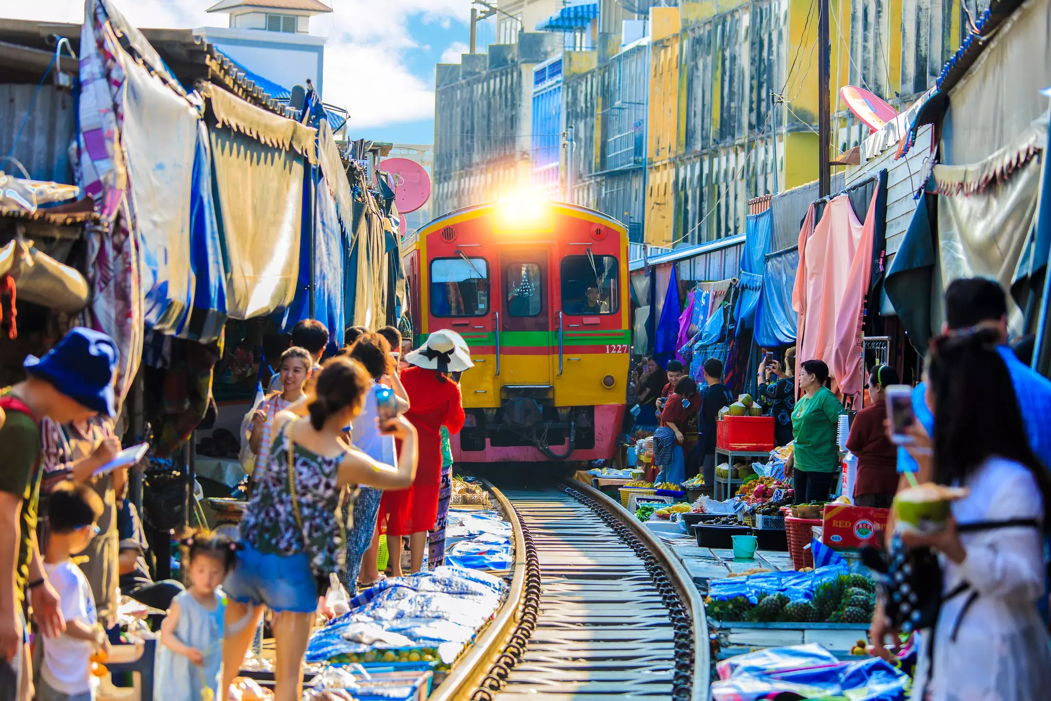 A train moves down a track surrounded by vendors and merchants, whose stalls and products almost touch the train. People lean out to take photos of the oncoming train.