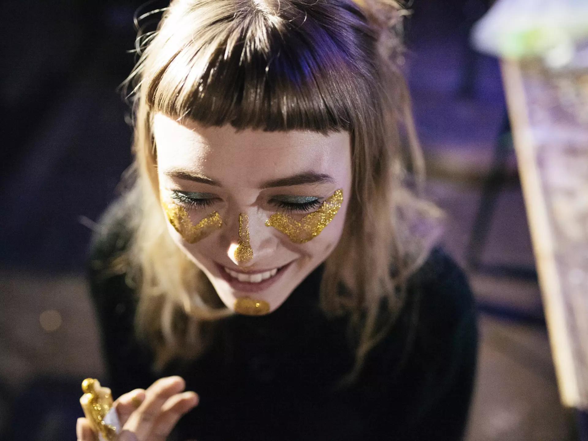A woman smiles as she paints her face with gold glitter while out clubbing