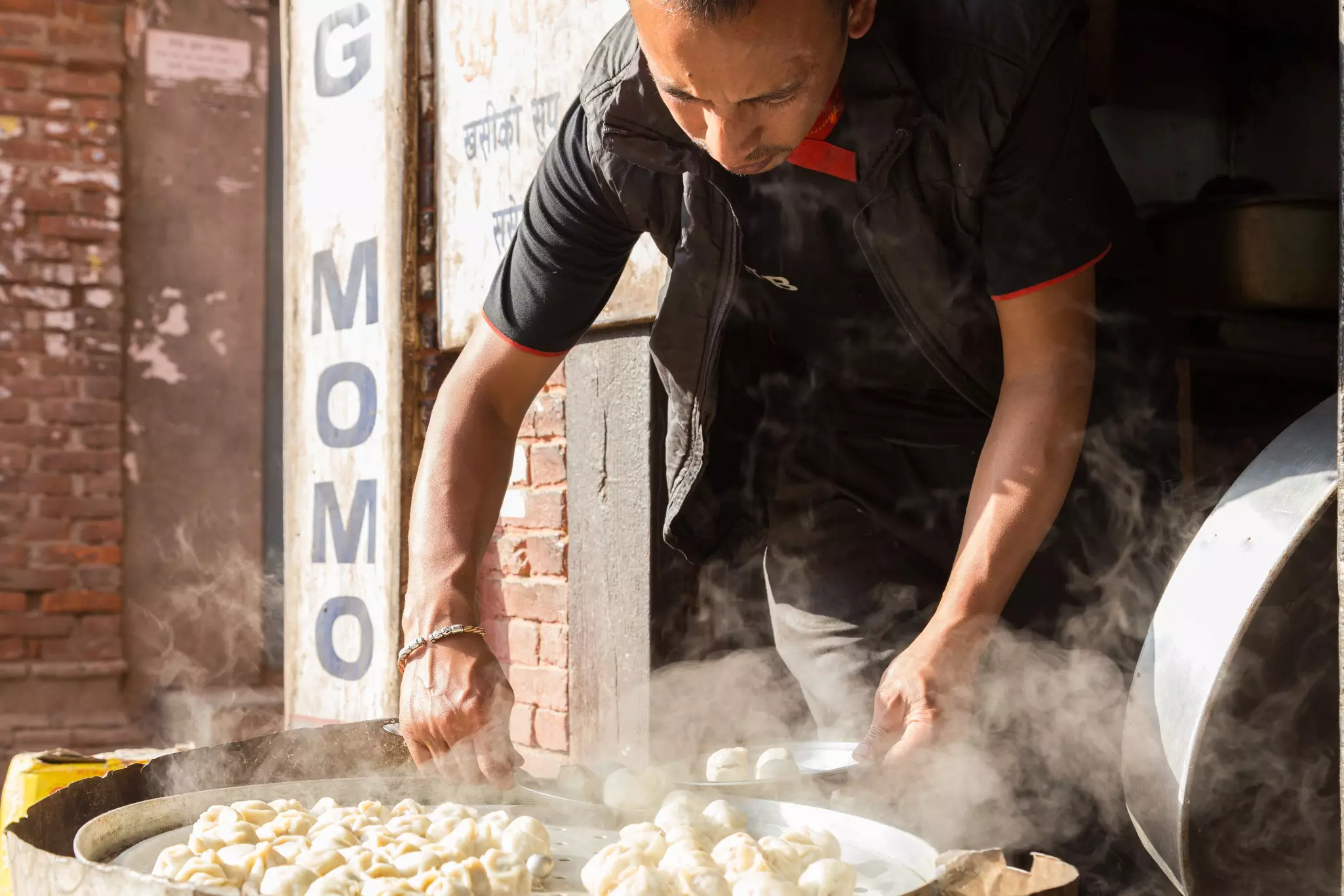 A man leans over a huge pot with dumplings at a streetside stall. Steam rises from the pot.