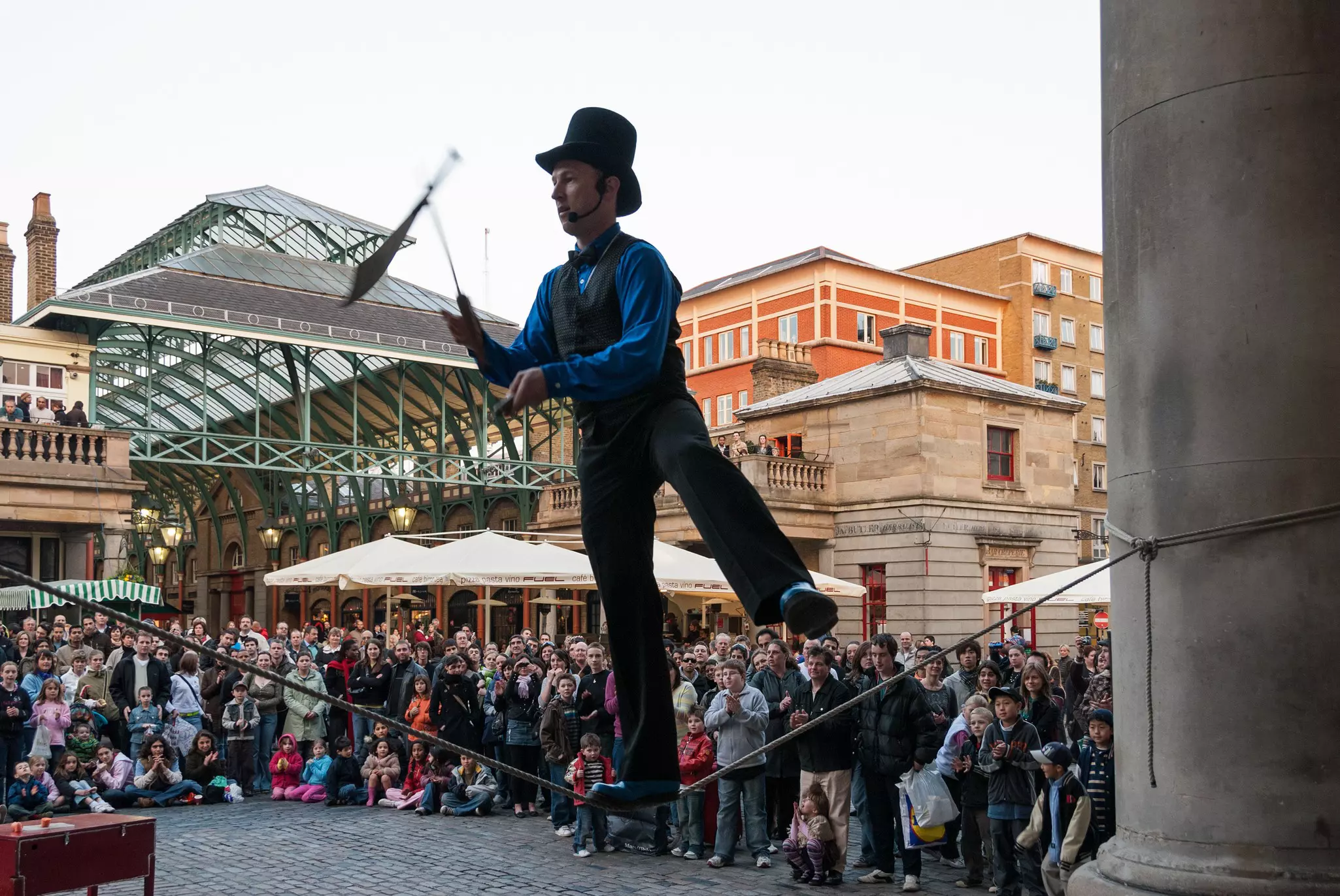 A street performer balances on a wire while juggling knives as a crowd looks on.