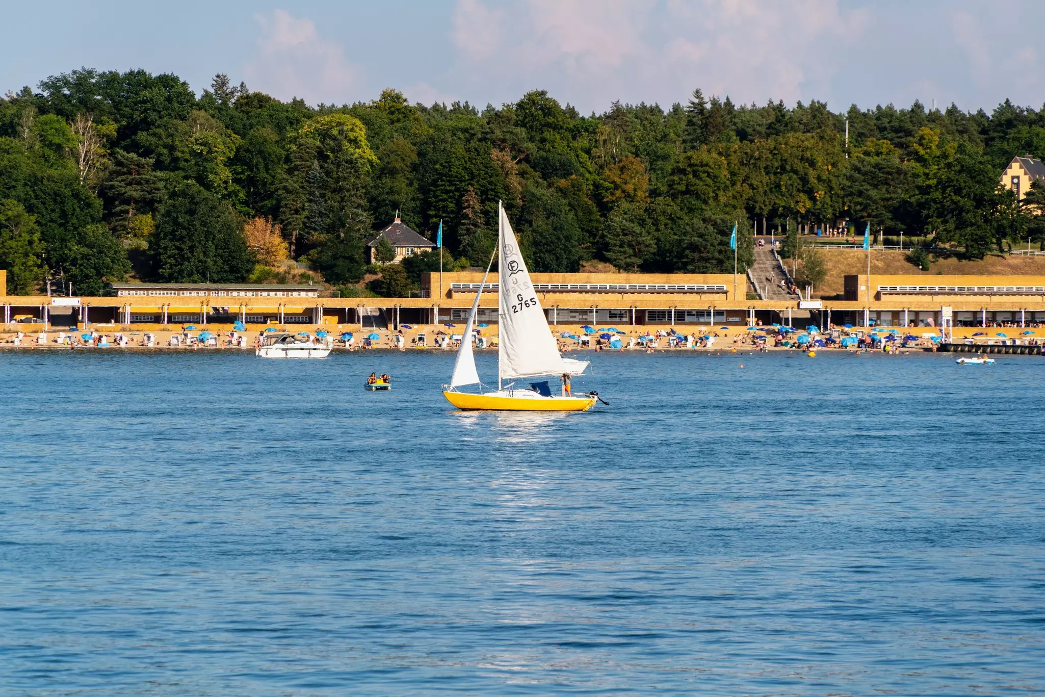 A sail boat in a lake as a peddle boat passes nearby. People lounge in the sun at the edge of the lake.