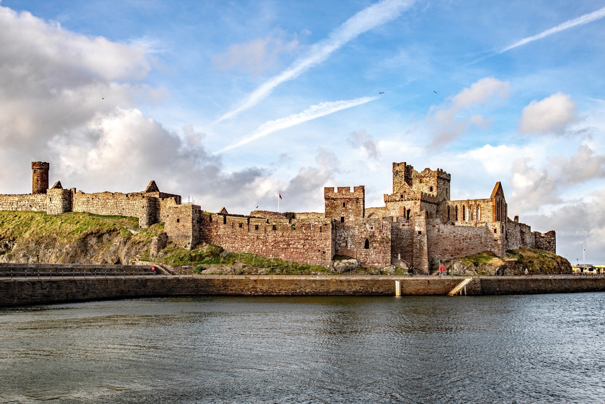 Ruined sandstone walls and towers of Peel Castle on St Patrick's Isle above calm water in Peel, Isle of Man