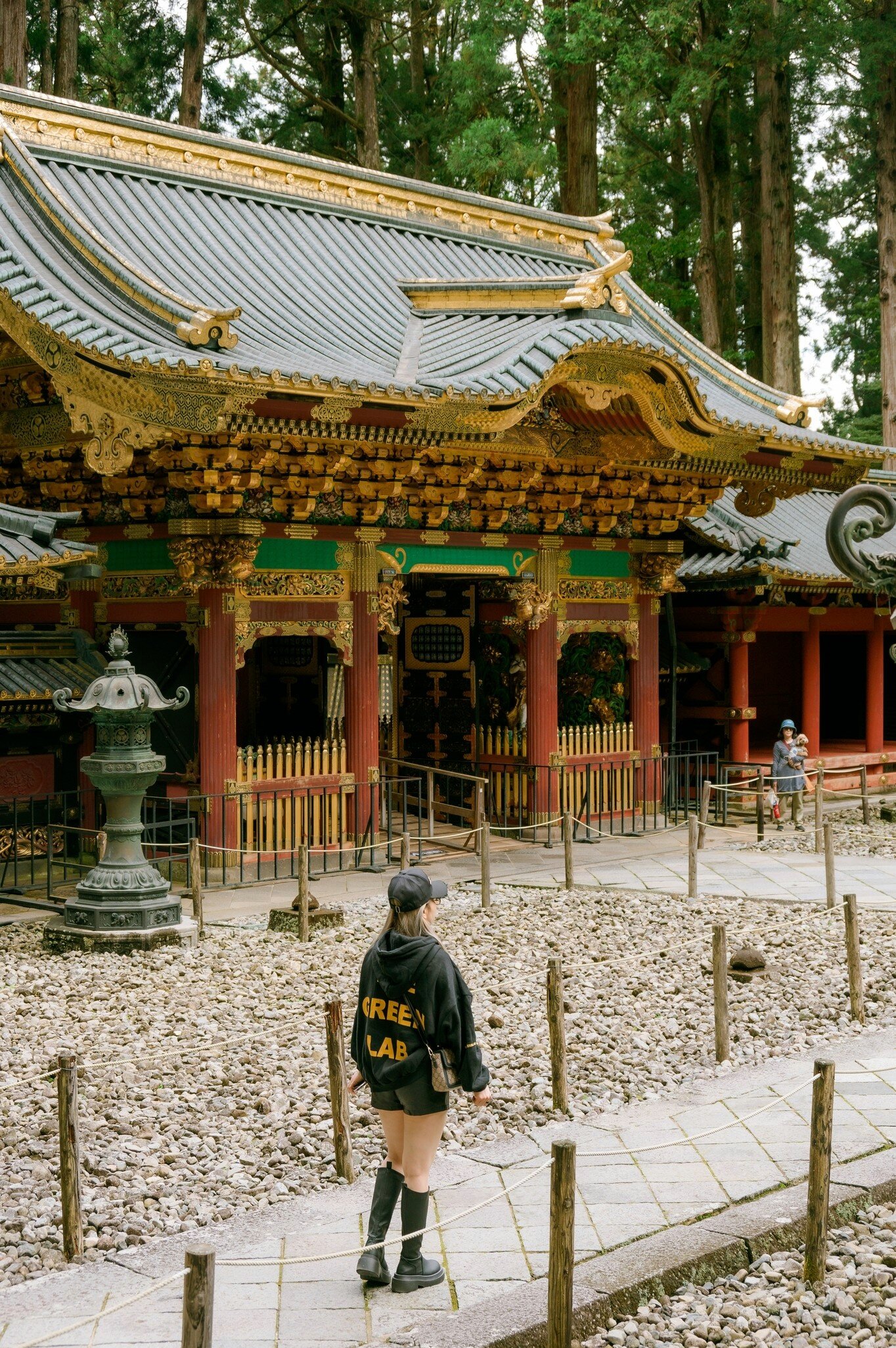 Visitor walks past the gilded, lacquered halls and stone lantern of Nikkō Tōshō-gū shrine, Japan; hoodie reads GREEN LAB