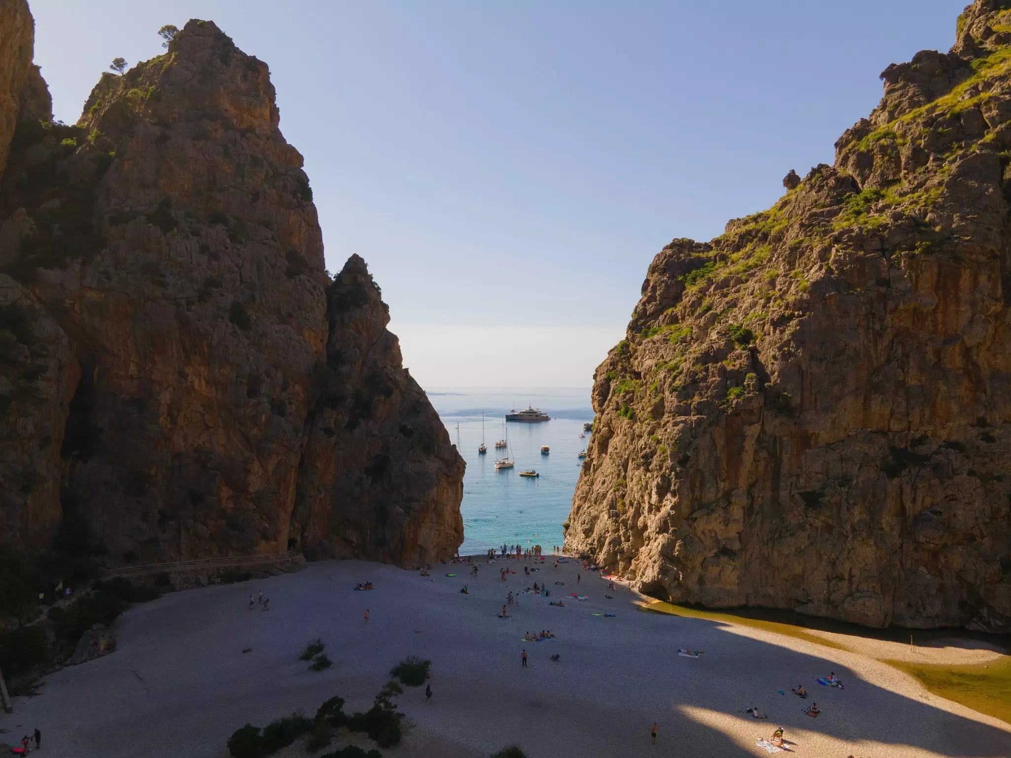 People on a sandy beach between two massive rocks that frame a boat in the water.