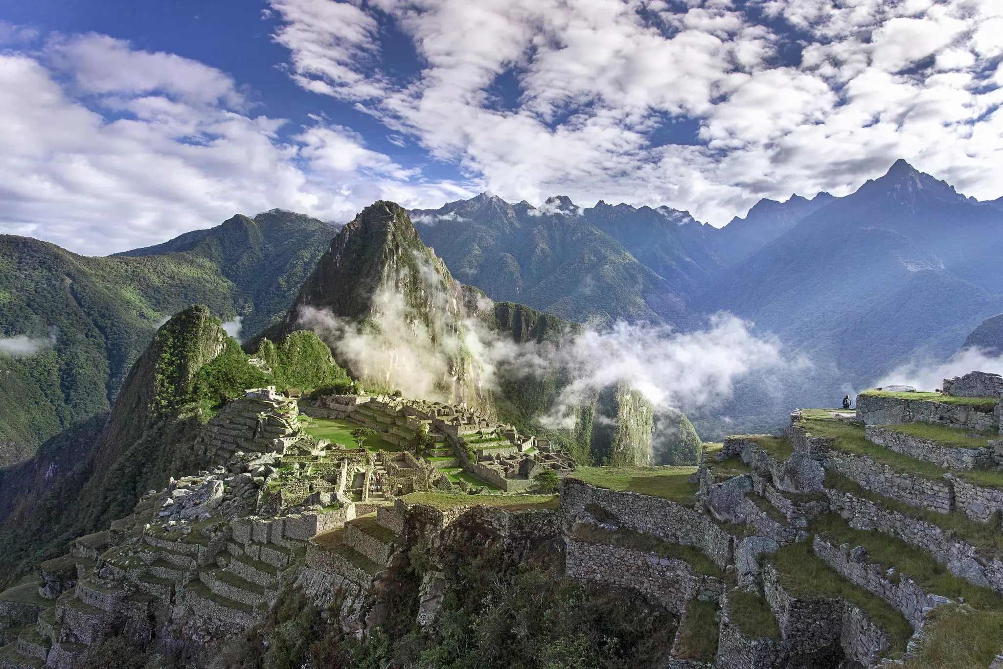 Ancient city walls built into tiers on steep rocky outcrops at high altitude, as wisps of cloud float over.