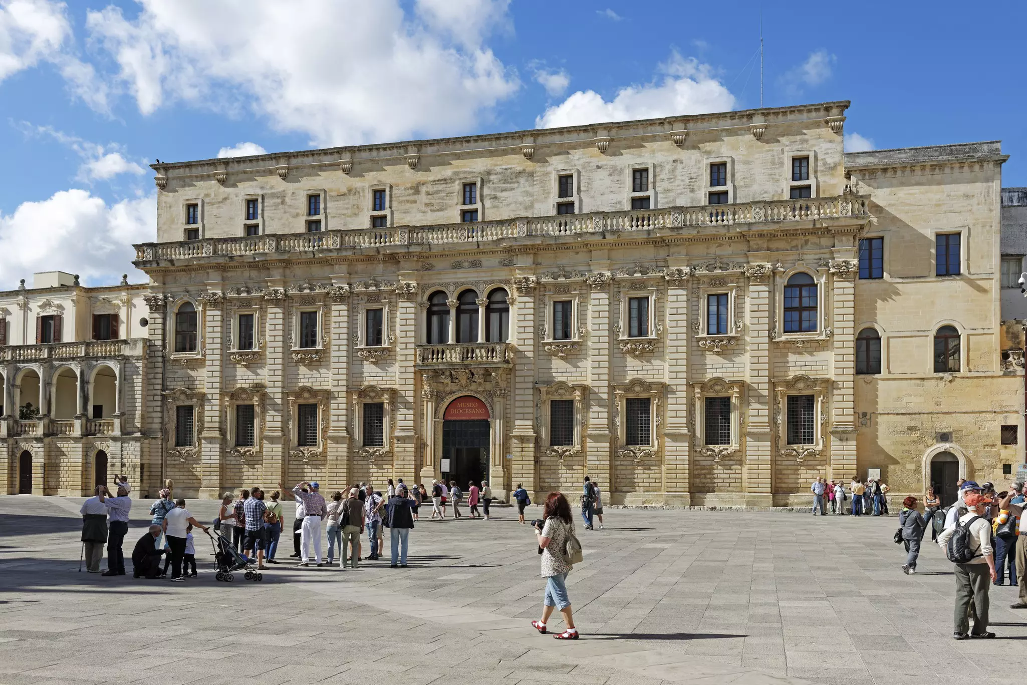 Visitors passing through a city square with a large grand mansion.