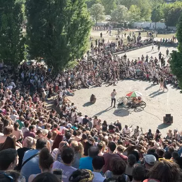 Thousands of people sit in an amphitheater in a city park watching a performer.