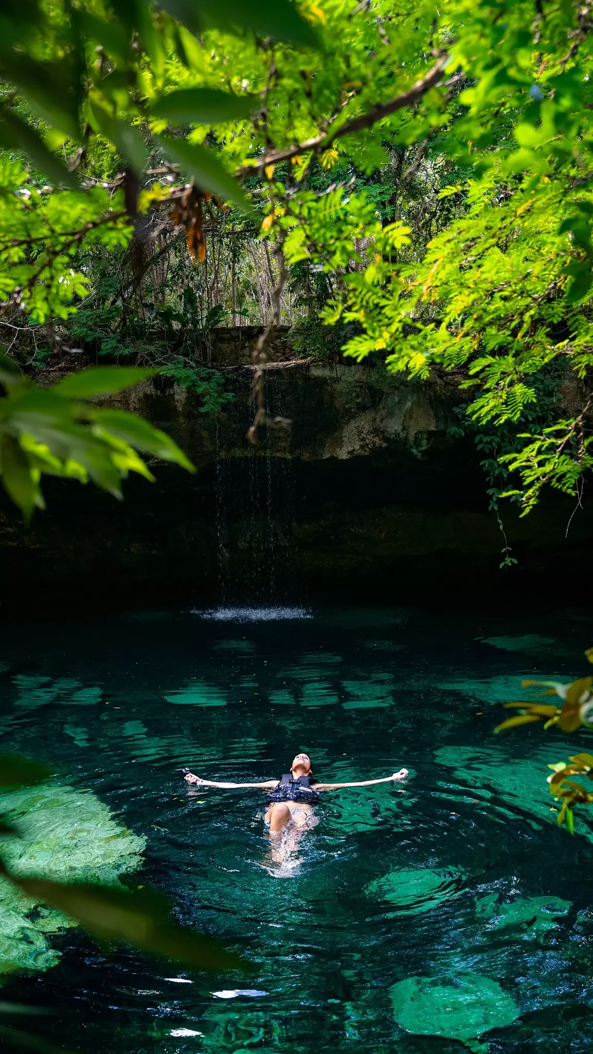 A woman floating in a tropical Mexican cenote with green turquoise crystal clear water surrounded by foliage, cliffs, and a small waterfall