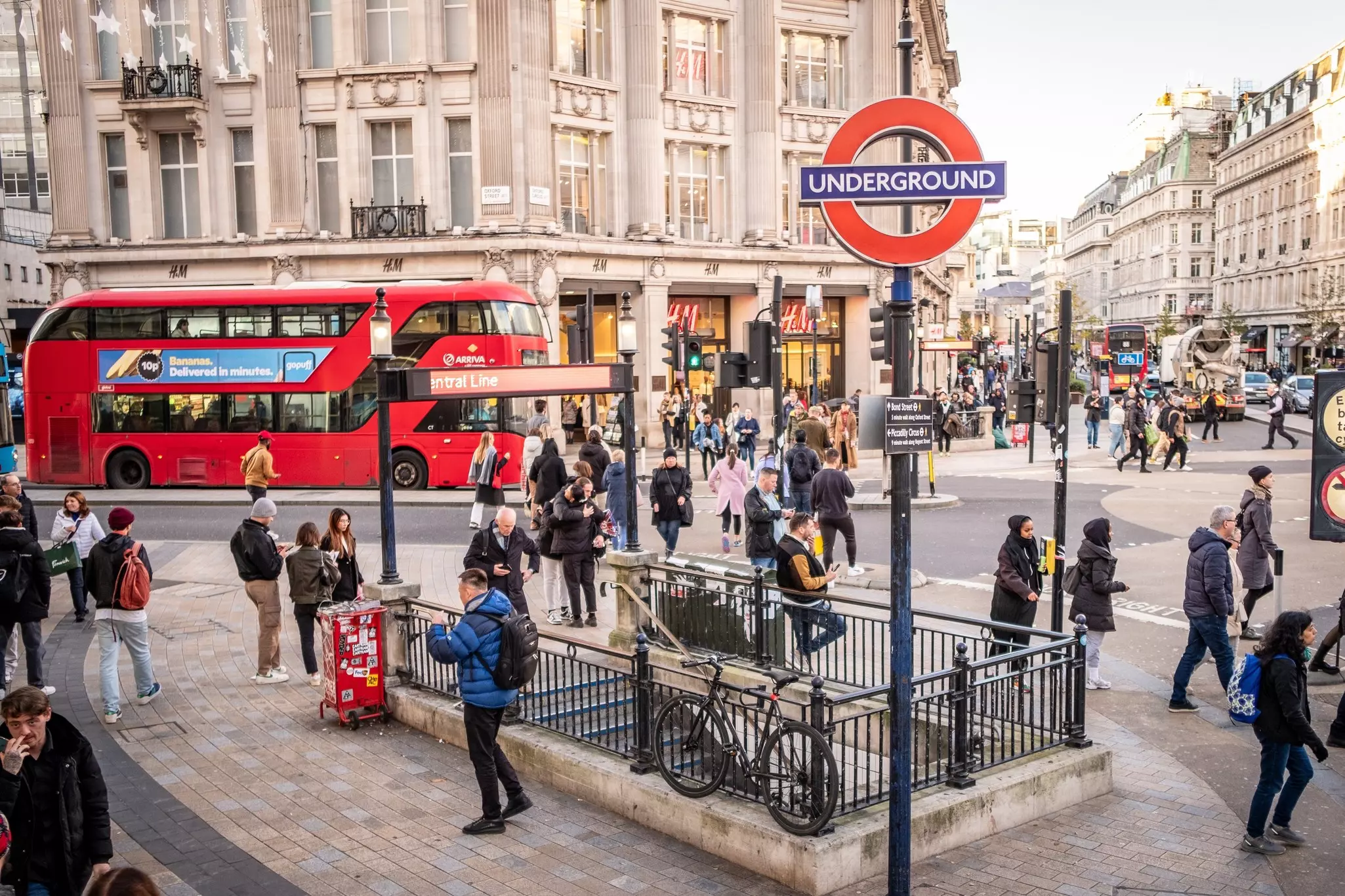 A road intersection in a busy city, with a red double-decker bus passing a round sign that says "Underground"