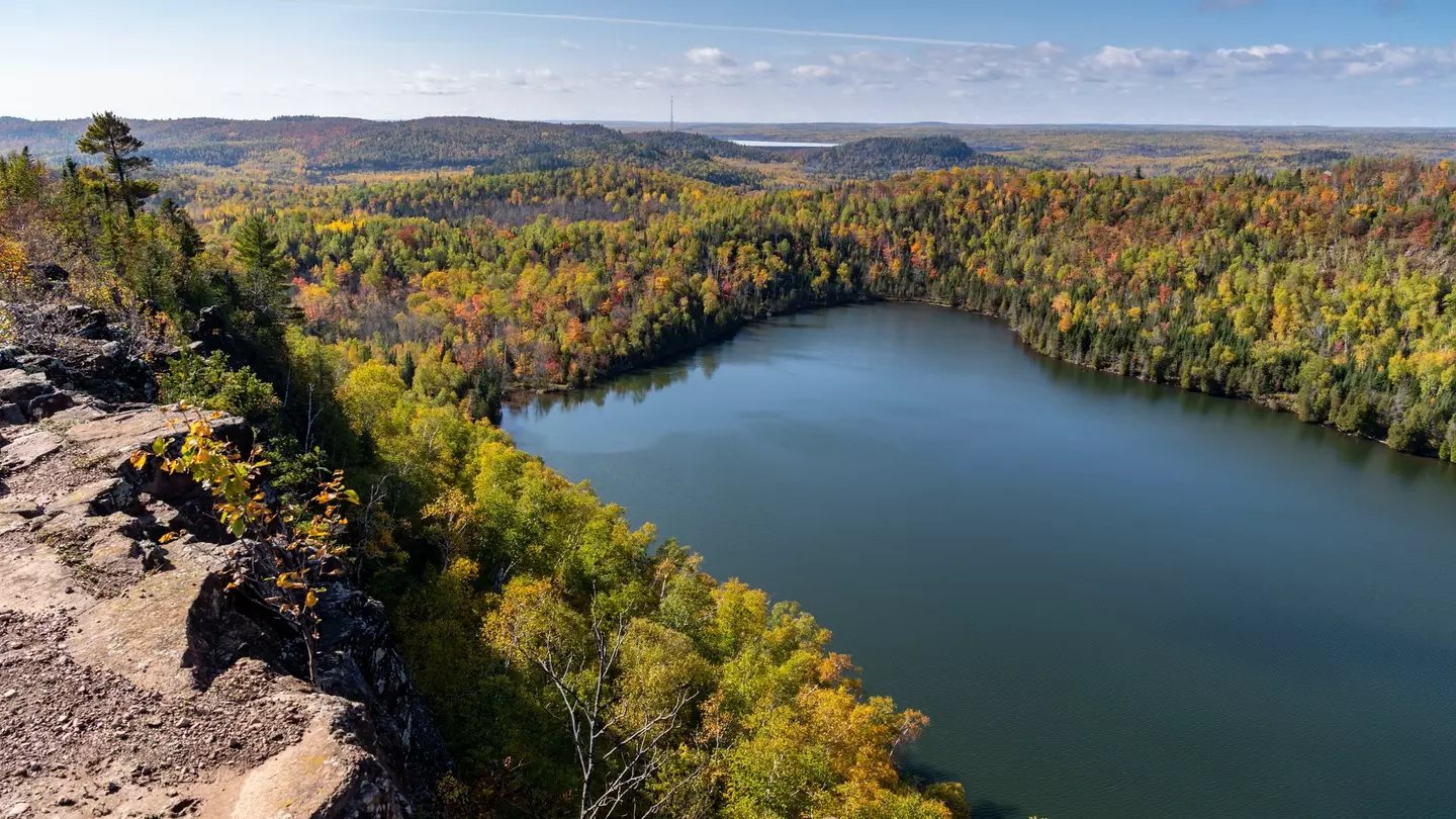 From a rocky outcrop, trees with some autumn color descend to the shore of a calm lake; the forest continues to the horizon, and another lake is in the far distance.