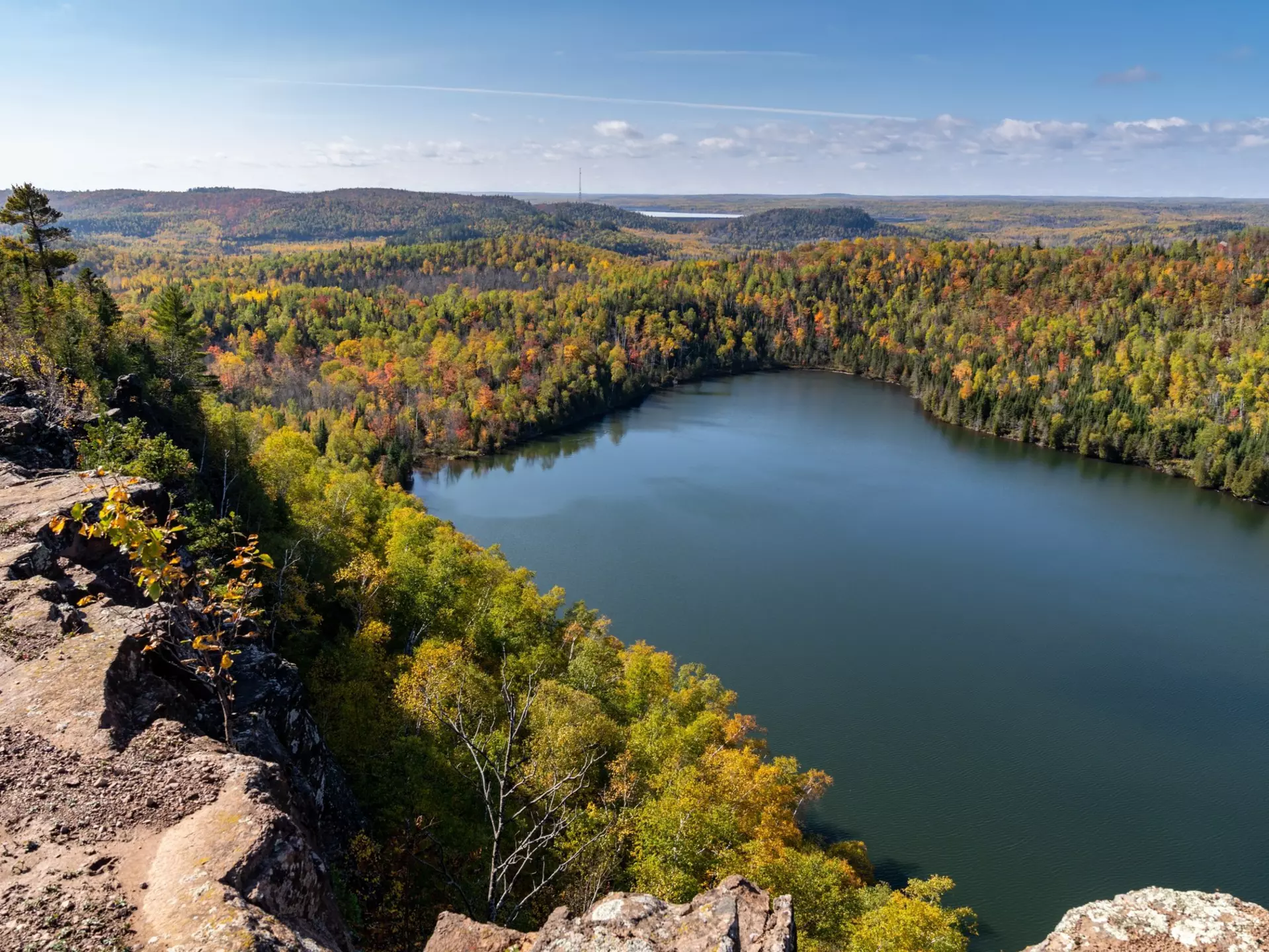 From a rocky outcrop, trees with some autumn color descend to the shore of a calm lake; the forest continues to the horizon, and another lake is in the far distance.