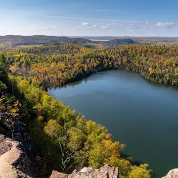 From a rocky outcrop, trees with some autumn color descend to the shore of a calm lake; the forest continues to the horizon, and another lake is in the far distance.