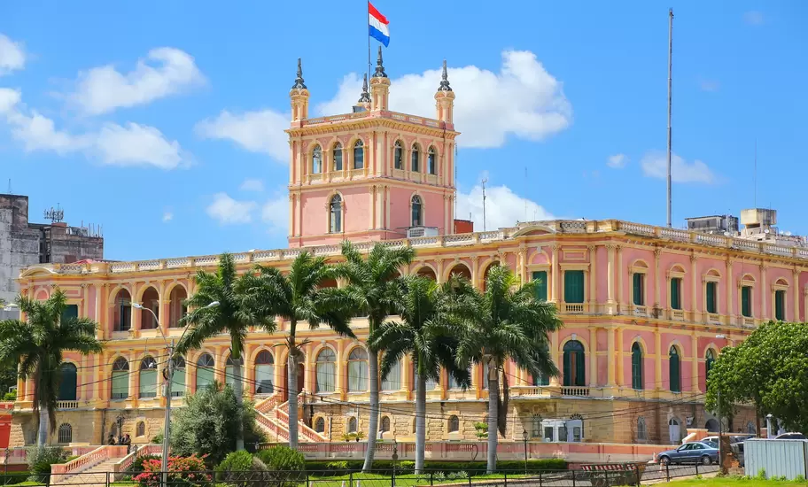 Pink and yellow Palacio de los López in Asunción, Paraguay, flying the Paraguayan flag behind palm trees and a lawn