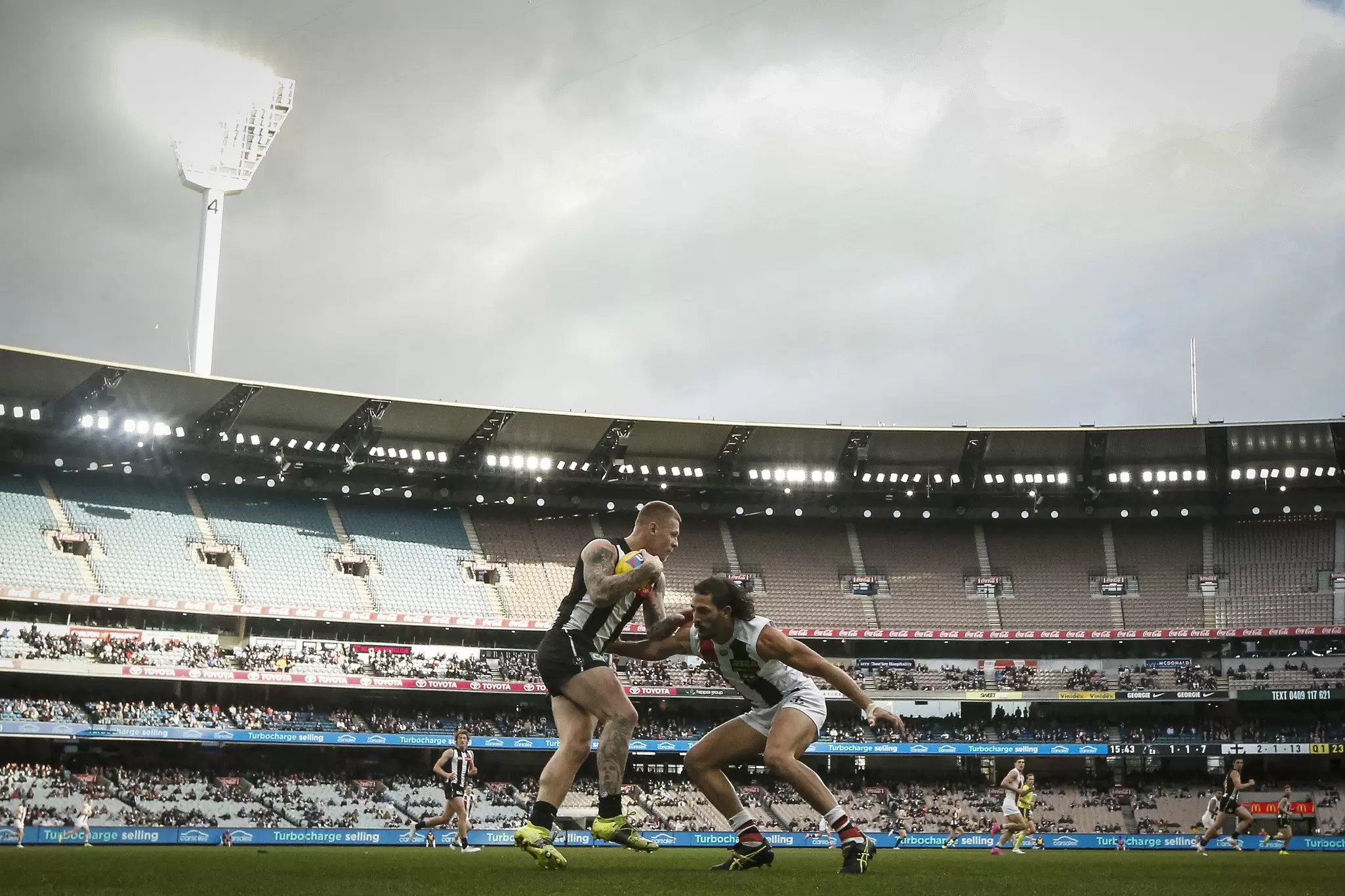 Two players tussle on the pitch of Melbourne Cricket ground during an AFL match between Collingwood Magpies and St Kilda Saints. The large grandstand of the stadium is visible in the background, with many fans watching on.