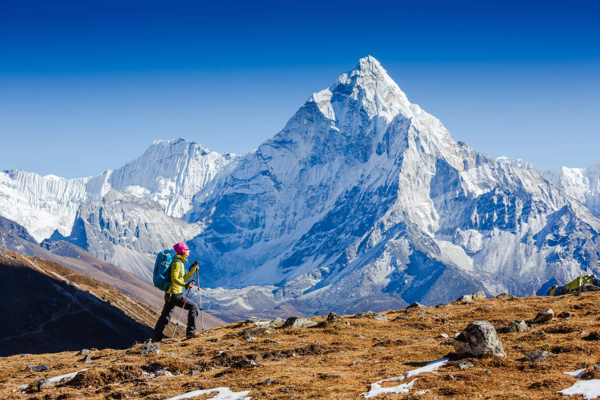 A trekker with poles navigates a sloping mountainside, with a huge, snow-covered peak visible across the valley.