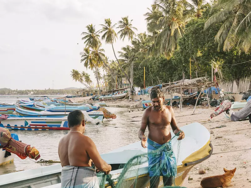 Two men inspect fishing nets by a boat on the sand.