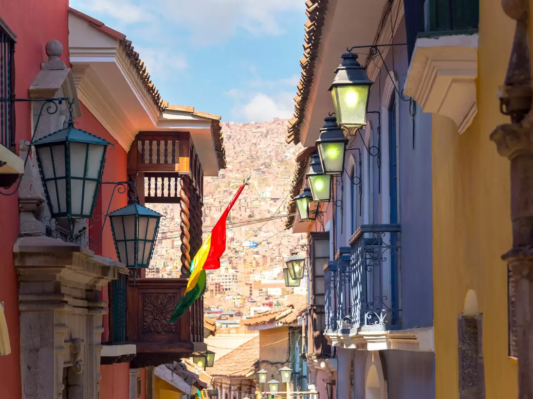 A narrow colonial street of red, blue and yellow facades hung with iron lanterns and carved wooden balconies, dense hillside housing filling the gap beyond.