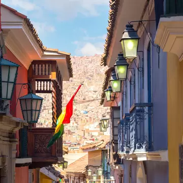 A narrow colonial street of red, blue and yellow facades hung with iron lanterns and carved wooden balconies, dense hillside housing filling the gap beyond.