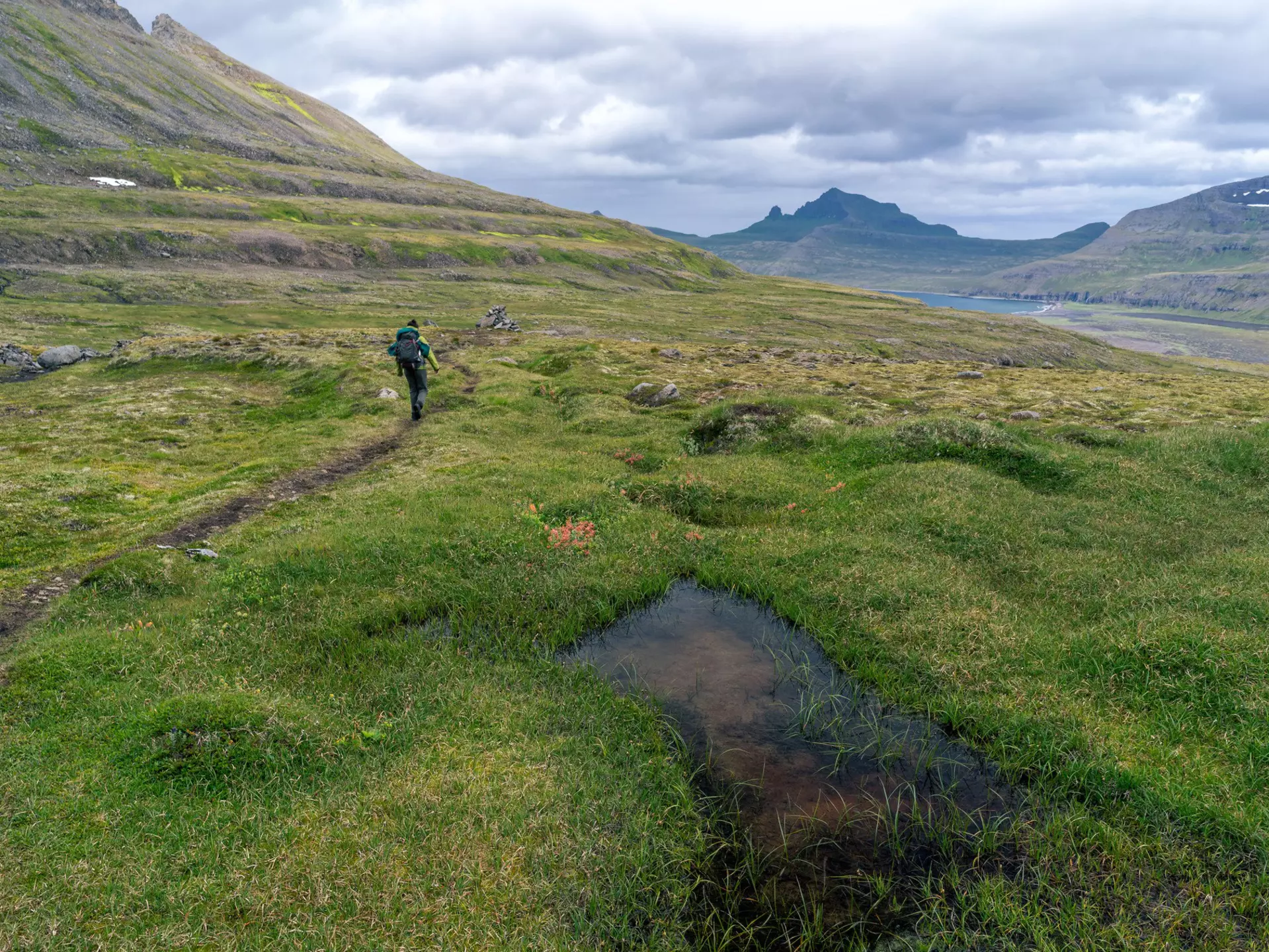 A hiker on a narrow path through a grassy valley, with clouds overhead