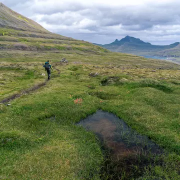 A hiker on a narrow path through a grassy valley, with clouds overhead