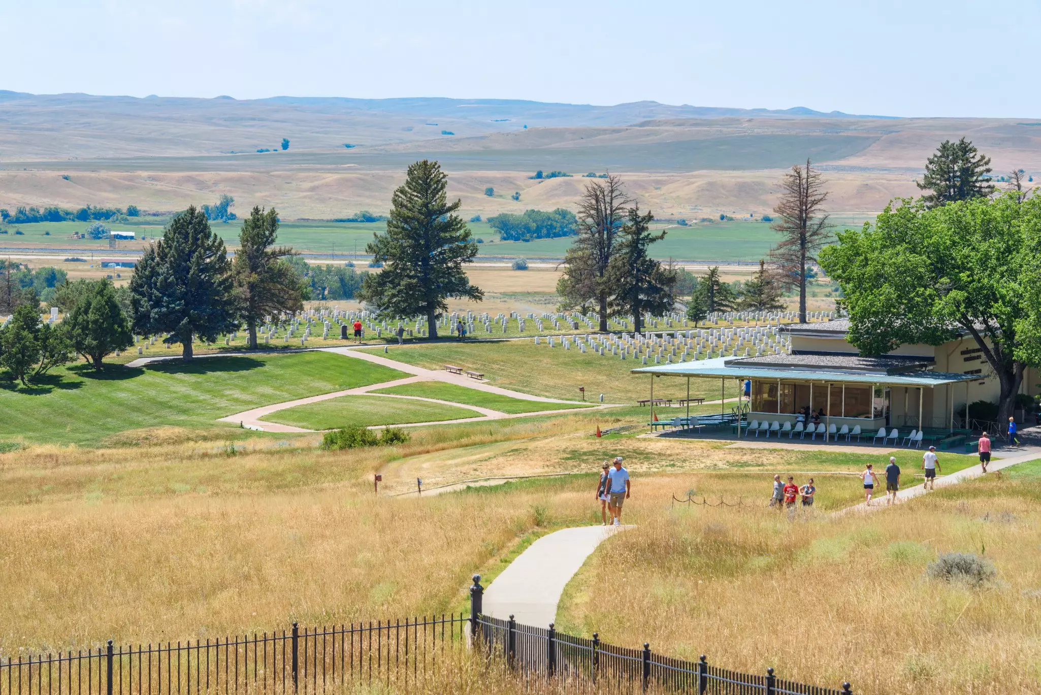 People follow a pathway near an interpretive center adjacent to a battlefield and graveyard marked with matching white headstones.
