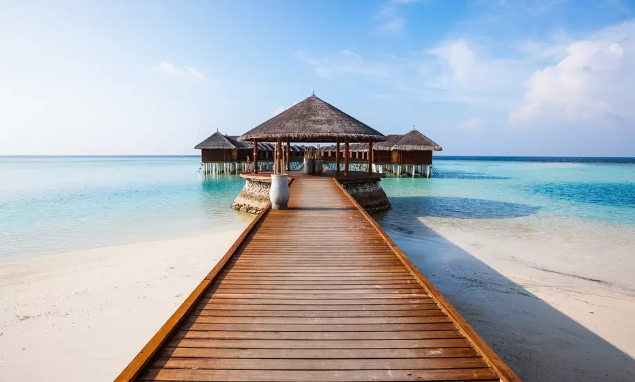 Maldives: wooden jetty crossing white sand to a thatched pavilion and overwater villas in a clear turquoise lagoon