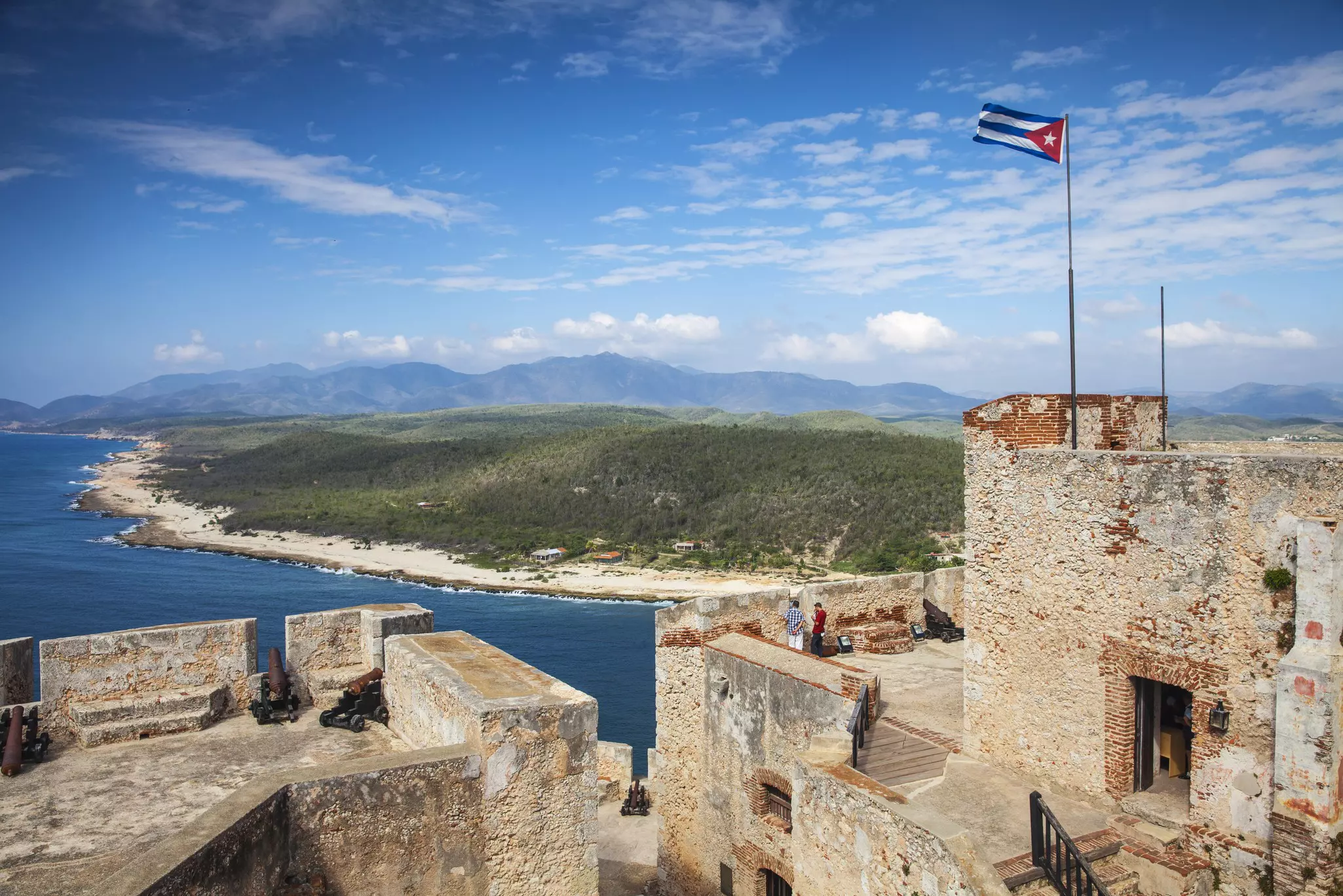 People stand on the top of a coastal fort looking out at the undulating coastline. A Cuban flag flies from the main fort tower.