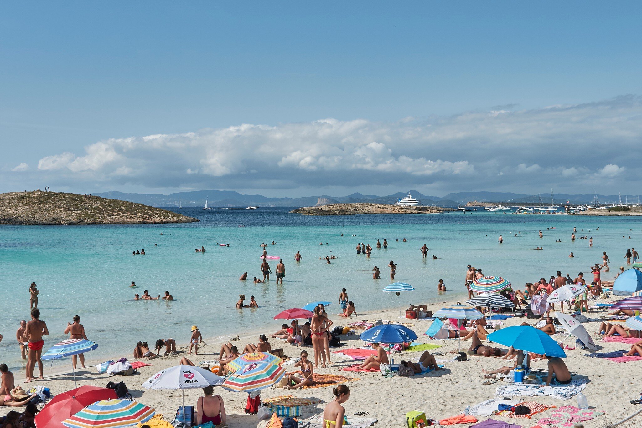 People under umbrellas relax on the sand of a large beach lapped by turquoise waves.